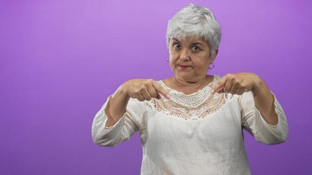 Woman with grey hair and hoop earrings pointing index fingers down at white lace blouse in studio with purple backdrop and direct gaze; assertive instruction.