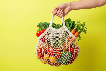 Fototapeta premium Hand holding reusable mesh bag with fresh vegetables on yellow background, eco friendly grocery and healthy lifestyle concept.
