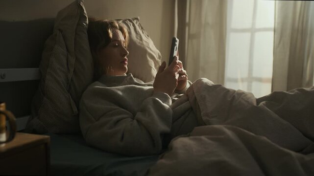 Stab shot of sleepless young Caucasian girl scrolling on smartphone and feeling bored while lying in bedroom with open window in low light