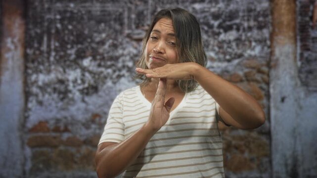 Woman making timeout hand gesture at a weathered building wall, wearing striped shirt, neutral expression; assertive dismissal.