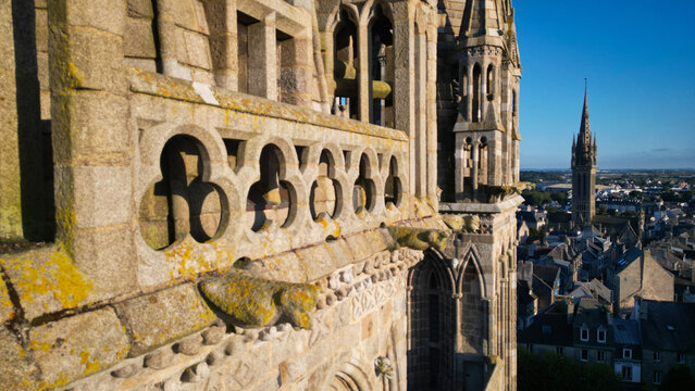 Aerial view of aged stone architecture, with intricate carvings and a distant spire piercing the horizon, bathed in the warm glow of the setting sun, Saint-Pol-de-Leon, France.