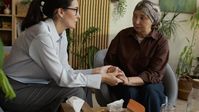 Tilt up slowmo shot of empathetic middle-aged Caucasian female psychologist holding mature Asian woman wearing headscarf by hands and supporting her during cancer counseling session in office