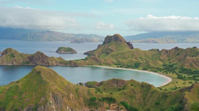Wide landscape view of Padar Hills overlooking calm turquoise bays in Komodo National Park, Flores, Indonesia. Rolling grassy hills and distant mountains create a dramatic tropical island panorama.