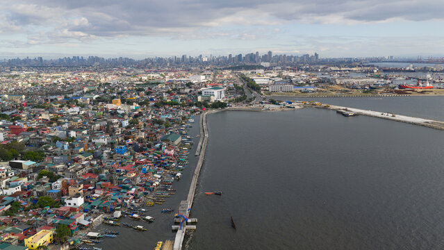 Aerial view of a coastal city where colorful houses meet the sea, juxtaposed against a distant skyline under a muted sky, Navotas, Metro Manila, Philippines.