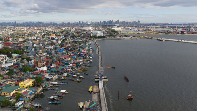 Aerial view of a bustling, colorful cityscape meeting the serene expanse of the sea, split by a narrow concrete path, Navotas, Metro Manila, Philippines.