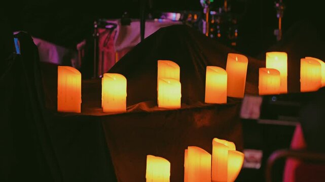 A cozy, atmospheric stage at a candlelit concert. Rows of glowing LED candles create a warm yellow glow against a dark background. A drummer plays the drums in the background