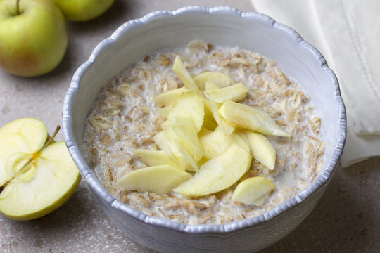 Oatmeal porridge with milk topped with fresh apple slices in ceramic bowl. Nutritious breakfast with oats and fruit on light kitchen table