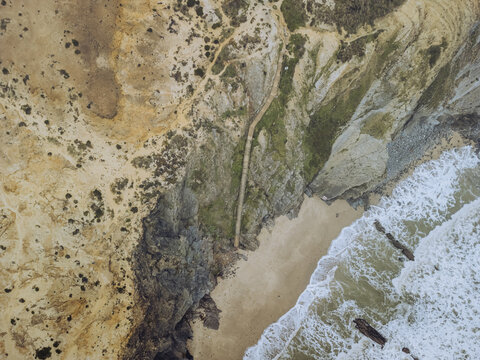 Aerial view of cliffs and foamy waves crashing on the sandy shore, contrasting with the earthy tones of the rugged landscape, Zambujeira do Mar, Portugal.