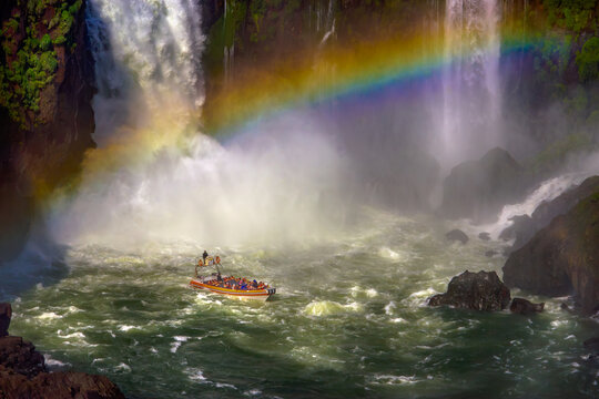 Speedboat full of tourists approaching powerful waterfalls with a rainbow at Iguazu Falls National Park in Argentina.