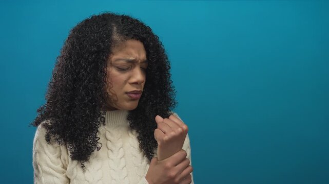 Woman in cream cable knit sweater clutching wrist with furrowed brow under soft lighting in blue studio; discomfort.