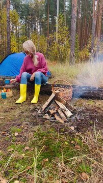 a blonde woman sits by a campfire next to a blue tent in a forest with golden autumn leaves, holding a mug in her hands and enjoying the beauty of the surrounding nature, her head lifted, her face rad