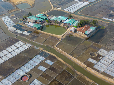 Aerial view of a village nestled amongst the geometric patterns of salt farms, with the tranquil pond reflecting the sky above, Cox's Bazar, Chittagong Division, Bangladesh.