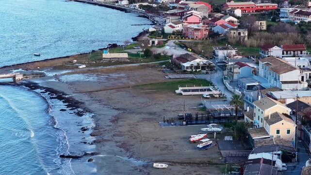 Drone footage over Roda village waterfront in Corfu Greece showing traditional houses fishing boats docked on sand and calm Ionian Sea at dusk