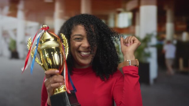 African american woman holds golden trophy with one hand and pumps raised fist beside her in outdoor building corridor; victory celebration.