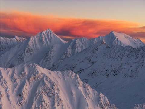 Aerial view of snow-capped peaks pierce the sky as the sun paints the clouds in fiery hues of orange and pink, Lyngen, Troms, Norway.