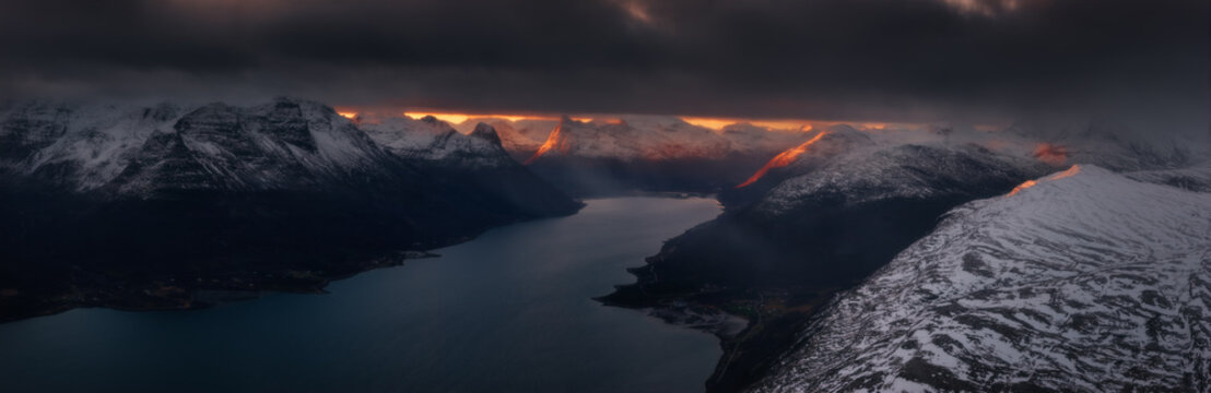 Aerial view of a fjord cutting through snow-dusted mountains, kissed by a dramatic sunset under a brooding sky, Lyngen, Troms, Norway.
