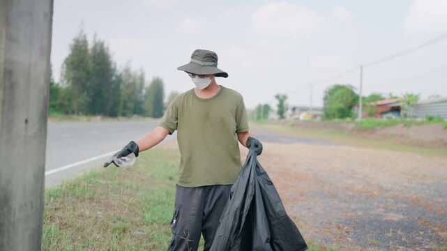 A dedicated Asian male environmentalist, fully equipped with protective gear, is diligently collecting litter along the roadside, demonstrating a spirit of environmental conservation and community res