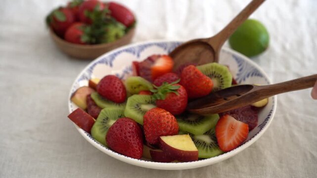 Hands mixing fresh fruit salad with strawberries, kiwi, apple, and orange in bowl, close up healthy homemade summer snack preparation concept