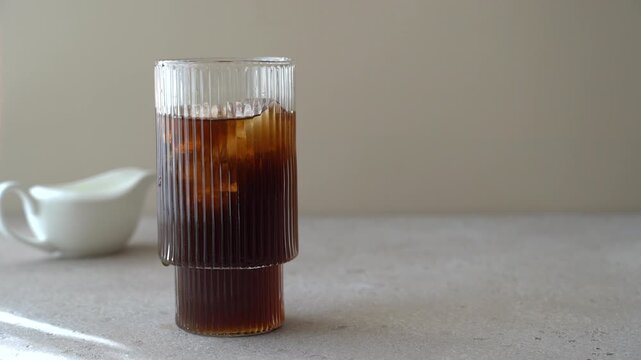 Female hand adding ice cubes into glass of black coffee on light background, close up iced coffee preparation, refreshing cold drink concept
