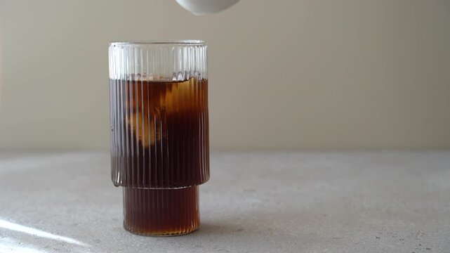 Milk pouring into glass of iced black coffee on light background, close up iced latte preparation, refreshing cold coffee drink concept