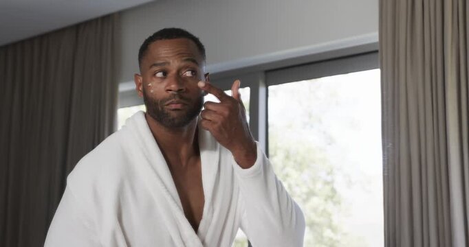 African American man dabbing and blending under-eye cream at bedroom window wearing white bathrobe