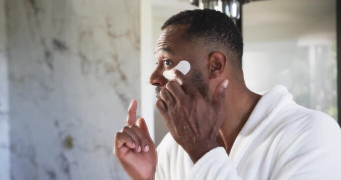 African American man standing at bathroom mirror wearing white bathrobe applying under-eye patches
