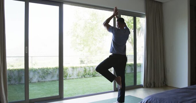 Mature man lifting foot, practicing tree pose on yoga mat by sliding glass door for balance