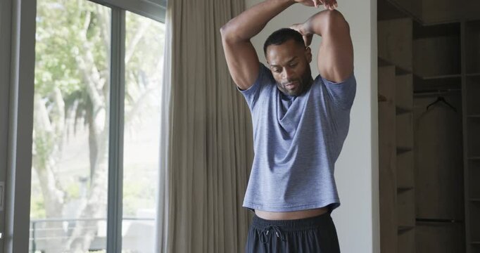 Mature man warming up cross-body to overhead triceps stretch by glass door, blue tee black shorts