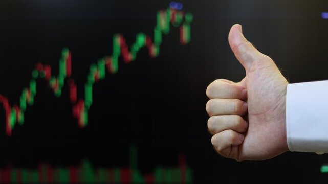 Close up of a male hand showing thumb up gesture against a stock market terminal. Businessman making approval sign in front of trading screen with candlestick chart. Financial success and profit conce