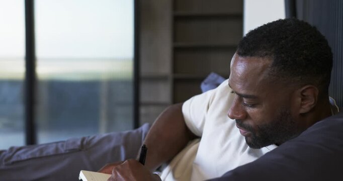 Mature African American man reclining on sofa by glass door, with poised pen journaling in notebook
