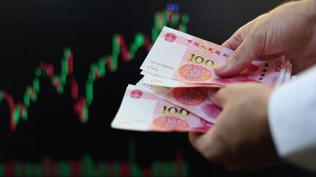 Close up view of man hands, Chinese Yuan banknotes, market chart on computer screen in background. Concept of currency growth, stock price change, volatile financial market, internet trading in China.