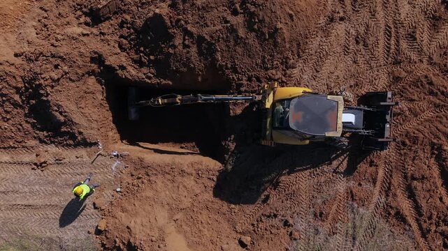 A top down aerial drone shot shows a backhoe loader excavating a deep trench while a construction engineer in safety gear supervises the earthworks on site.