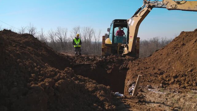A backhoe loader digs a deep trench at a construction site while a construction engineer in safety gear supervises the excavation from the edge of the earthworks area.