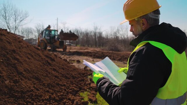 A construction engineer in safety gear reviews project plans while a backhoe loader operates in the background at an active earthmoving site.