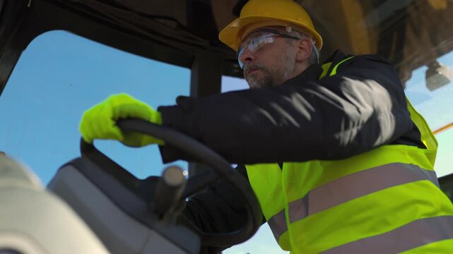 A construction worker in protective gear operates heavy machinery from the cabin while working at an outdoor job site.