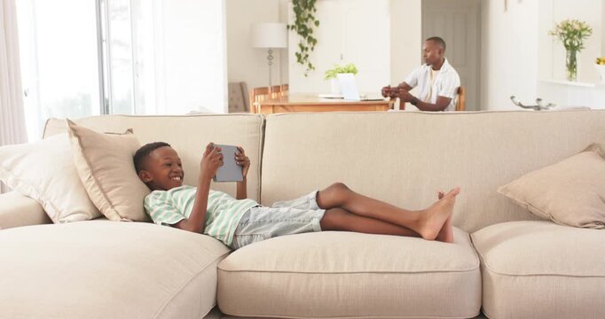 Diverse family child lying on sofa with tablet mother bringing mugs father using laptop looking up