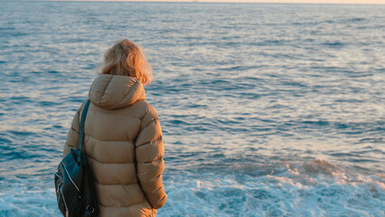 Woman in puffer jacket stands by ocean during sunset with waves crashing © Наталья Добровольска