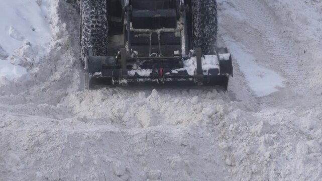Snowplow tractor scraping a dense layer of snow down to the asphalt. Close-up of a utility loader bucket clearing the road after a blizzard