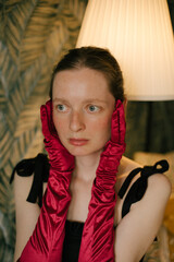 Elegant freckled woman in black dress and red gloves posing in vintage interior with soft lamp light.
