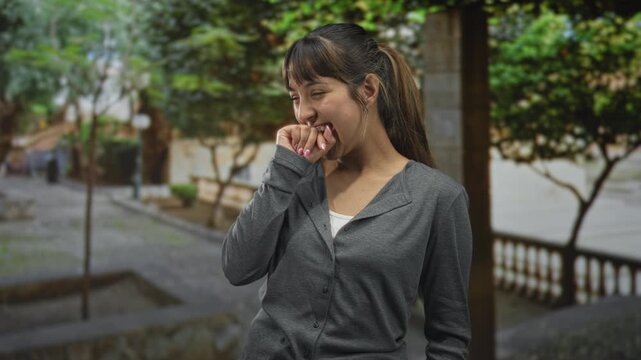 Young woman biting her hand and smiling, fingernails visible, wearing gray cardigan, standing on a street with trees and stone balustrade; playful shyness.