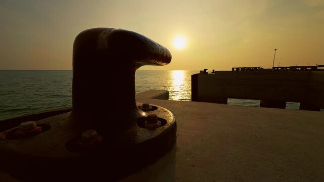 Massive Harbor Bollard Silhouette on Concrete Pier During Golden Sunset Over Calm Sea Horizon With Warm Light Reflection on Water. Maritime Infrastructure Navigation Shipping Logistics and Coastal Ind
