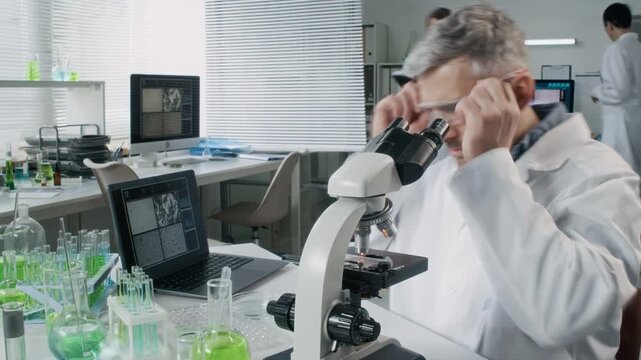 Caucasian senior man as male scientist looking in microscope and analyzing samples while sitting at table in lab with team in background, time lapse shot