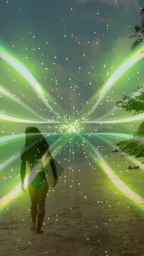 Vertical video: Surfer walking toward sea, green rays appearing on horizon over her guiding surf