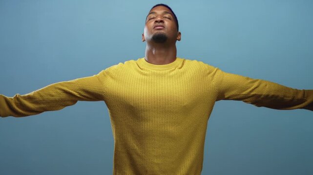 Black man with arms outstretched wearing a yellow sweater in studio, head tilted up and eyes closed in a relaxed breathing pose showing openness and release; freedom release breath.
