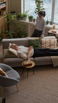 Vertical high angle shot of elderly Caucasian man lying on couch in modern therapist office and listening to middle-aged female psychologist conducting individual session