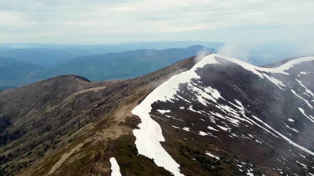 Aerial Mountain Ridge With Snow Sweeping Along Sharp Alpine Peak, Drone Reveals Layered Blue Ranges And Distant Horizon Under Overcast Sky, Crisp Rock And Patchy Snow Textures, Remote Wilderness Mood