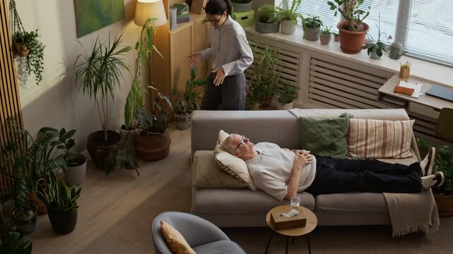 High angle shot of middle-aged Caucasian female therapist explaining psychological concept to mature male patient lying on couch, moving around green office while conducting individual consultation