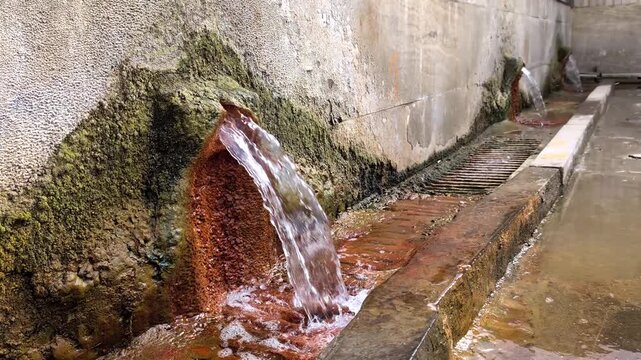 Ferruginous mineral spring water flowing from a stone fountain with rust colored deposits and green moss on a concrete wall showing its curative properties