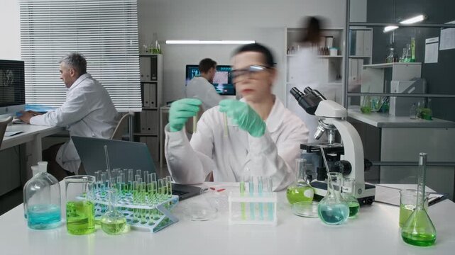 Time lapse shot of adult Caucasian woman as female scientist sitting at table in lab and doing tests, team in background, front view, copy space