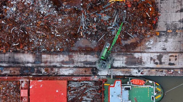 Close up aerial top view of scrap metal loading process. Green hydraulic excavator moving rusty metal scrap from dock pile into bulk carrier cargo hold.